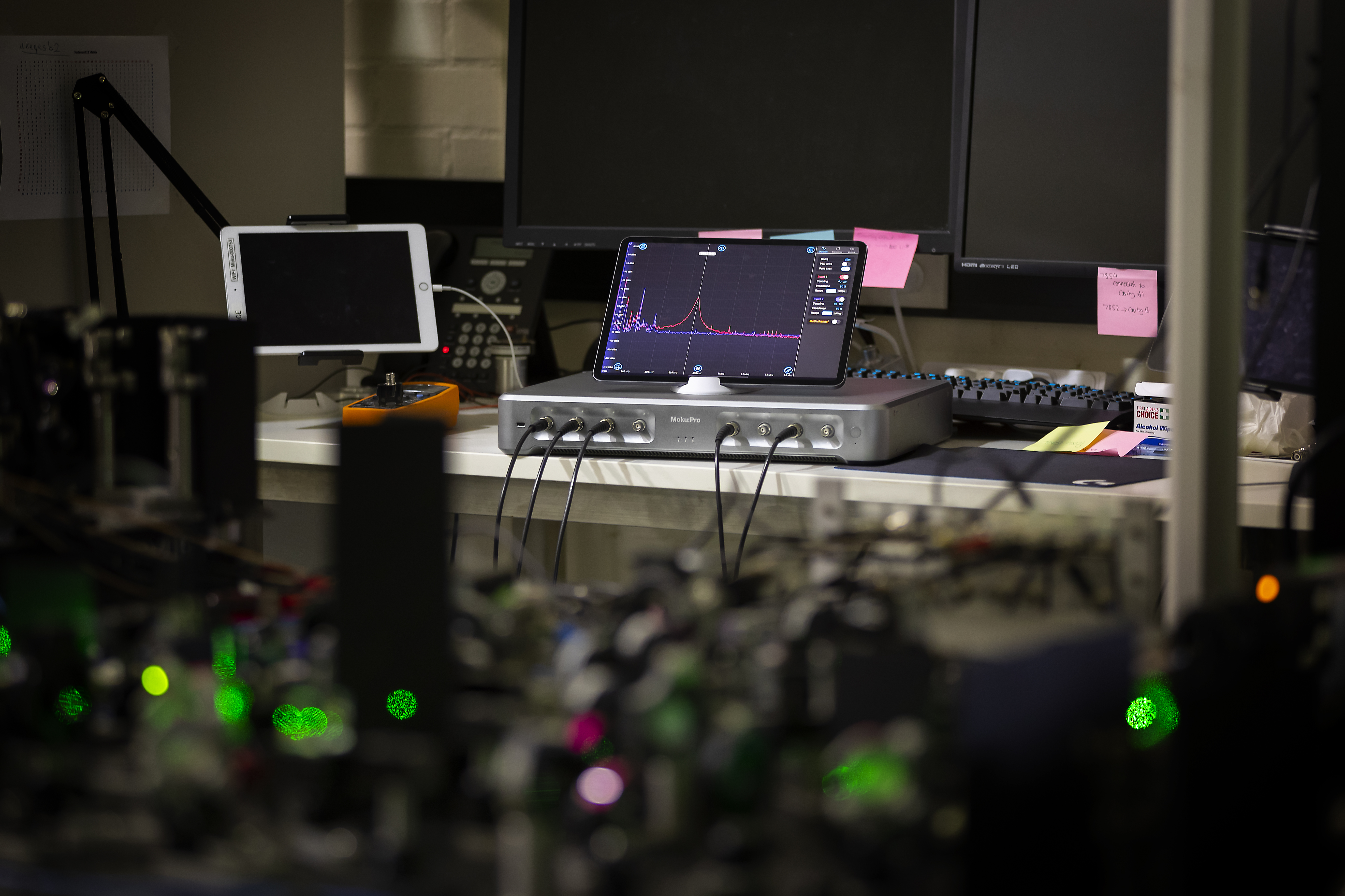 A Moku:Pro and iPad sit at a desktop workstation. In the foreground are optical components with a laser beam passing through them.  